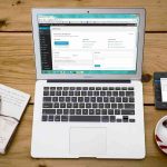 Laptop with open browser, smartphone, coffee cup, and notebook on wooden table.