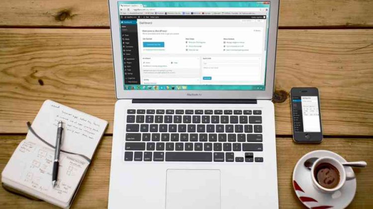 Laptop with open browser, smartphone, coffee cup, and notebook on wooden table.